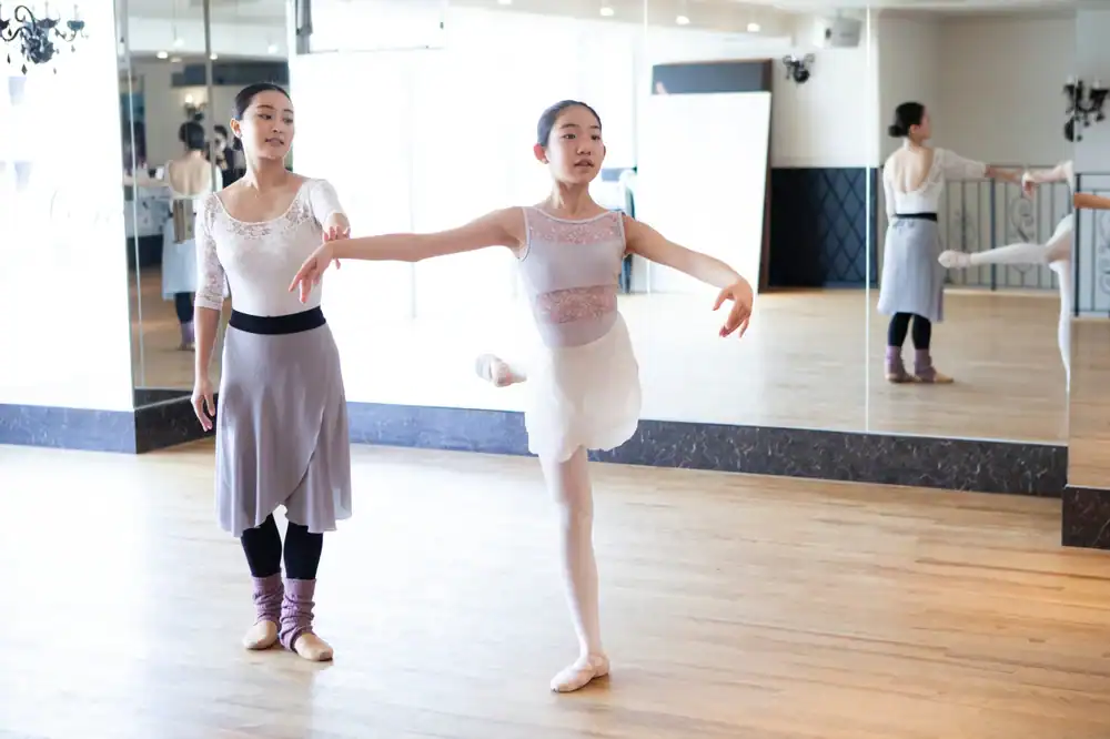 A ballet instructor watches a young student practicing in a studio with wooden floors and large mirrors, reminiscent of spaces used for ballroom dance Suffolk County, NY. The student balances on one leg, her arms gracefully extended.