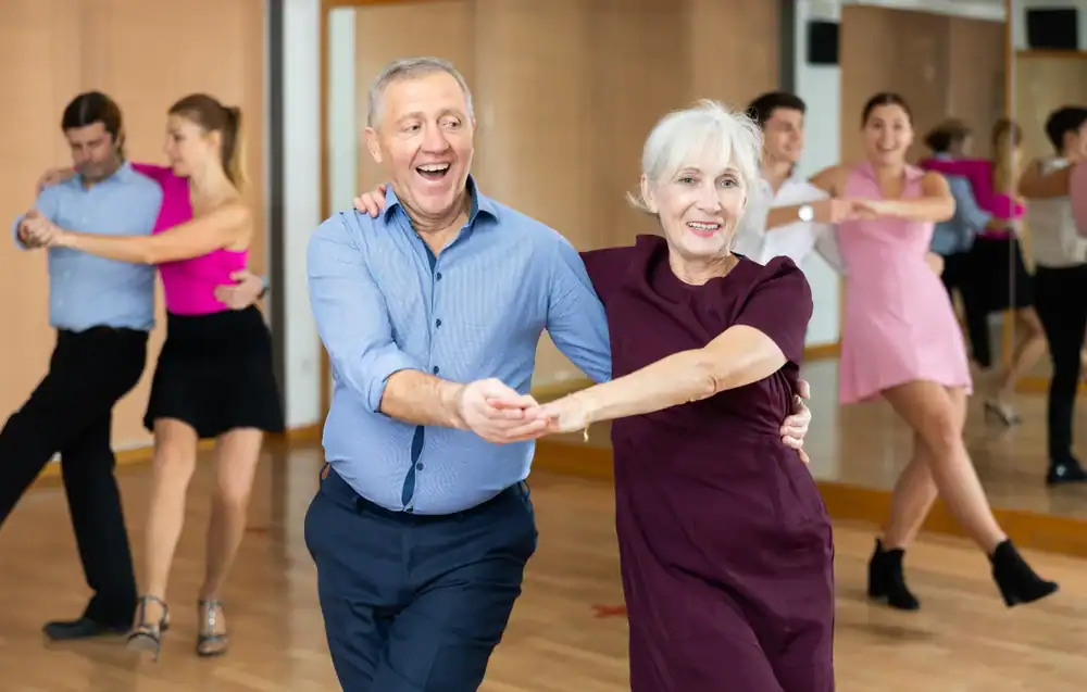 Older man and woman smiling and dancing together in a bright studio, with two other younger couples enjoying ballroom dance Suffolk County, making the most of their dance lessons in NY.