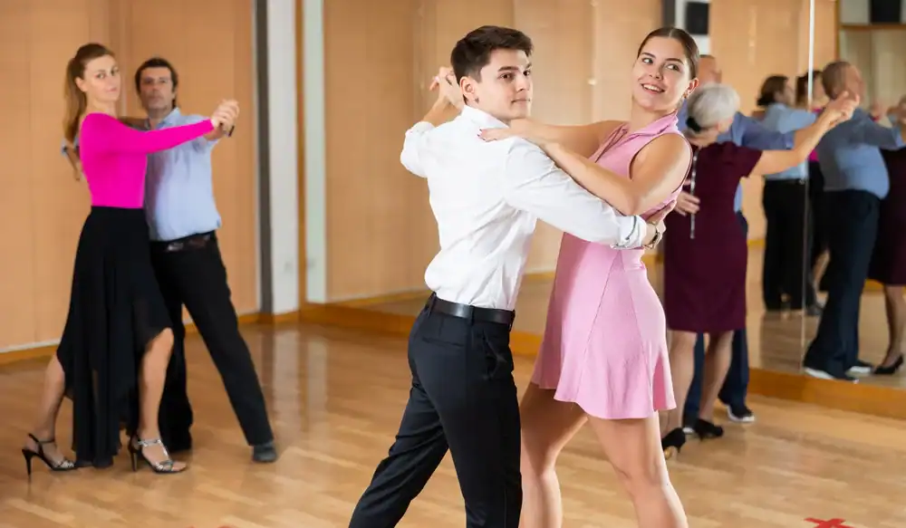 Two couples are ballroom dancing in a studio with wooden floors and mirrored walls. The smiling foreground couple wears light attire, while another duo glides behind&mdash;capturing the joy of ballroom dance in Suffolk County, NY.