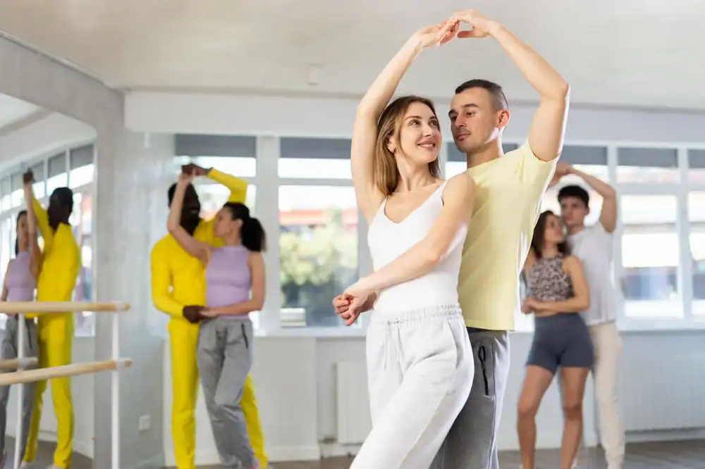 Several couples practice ballroom dancing in a bright studio with large windows and mirrors. In the foreground, a smiling woman and her partner, both in casual workout clothes, enjoy ballroom dance lessons in Suffolk County, NY.