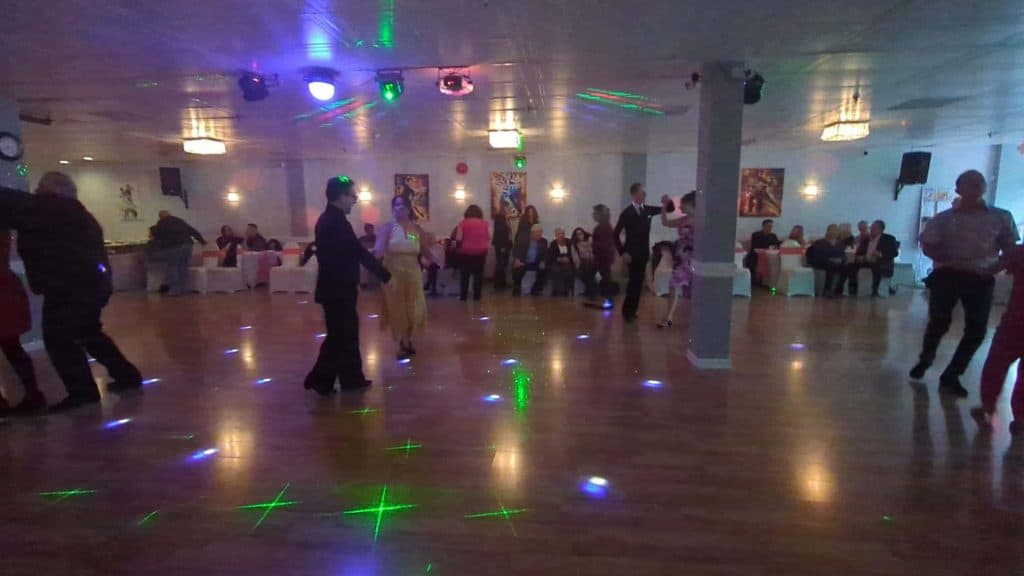 People enjoy ballroom dance in Suffolk County on a wooden floor in a dimly lit hall with colorful lights, while others sit along the walls watching. Chandeliers and wall art decorate this elegant space, perfect for Dance Lessons in Suffolk County, NY.