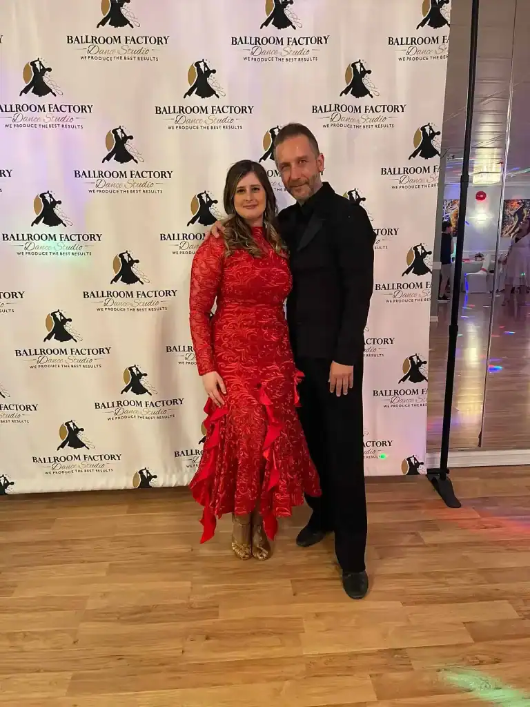 A woman in a red ruffled dress and a man in a black suit pose together on a wooden floor in front of a Ballroom Factory step-and-repeat banner, highlighting the elegance of ballroom dance Suffolk County, NY.
