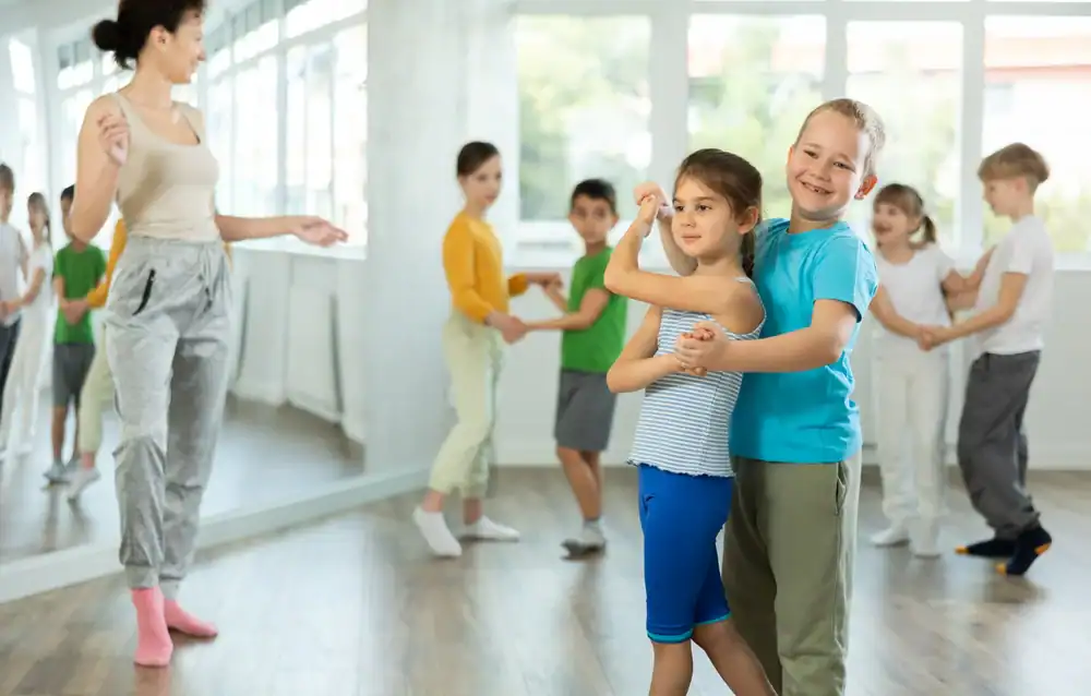 Children practice ballroom dancing in pairs in a bright studio, with a teacher guiding them. Large windows let in natural light as they enjoy their Dance Lessons Suffolk County, appearing happy and engaged throughout the lesson.