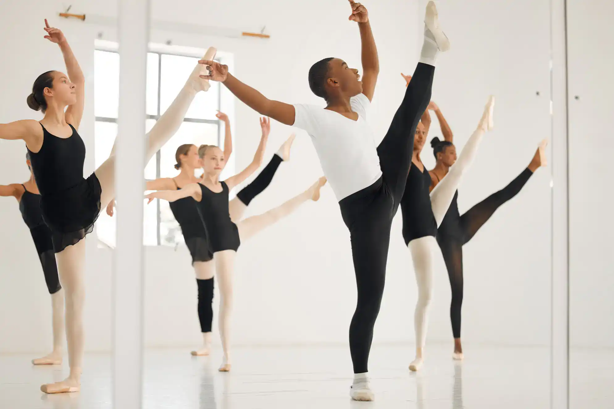 A group of young ballet dancers practice in a bright studio in Suffolk County, NY, performing high leg stretches at a barre. Focused and poised, they refine their form during dance lessons alongside others learning ballet and ballroom dance.
