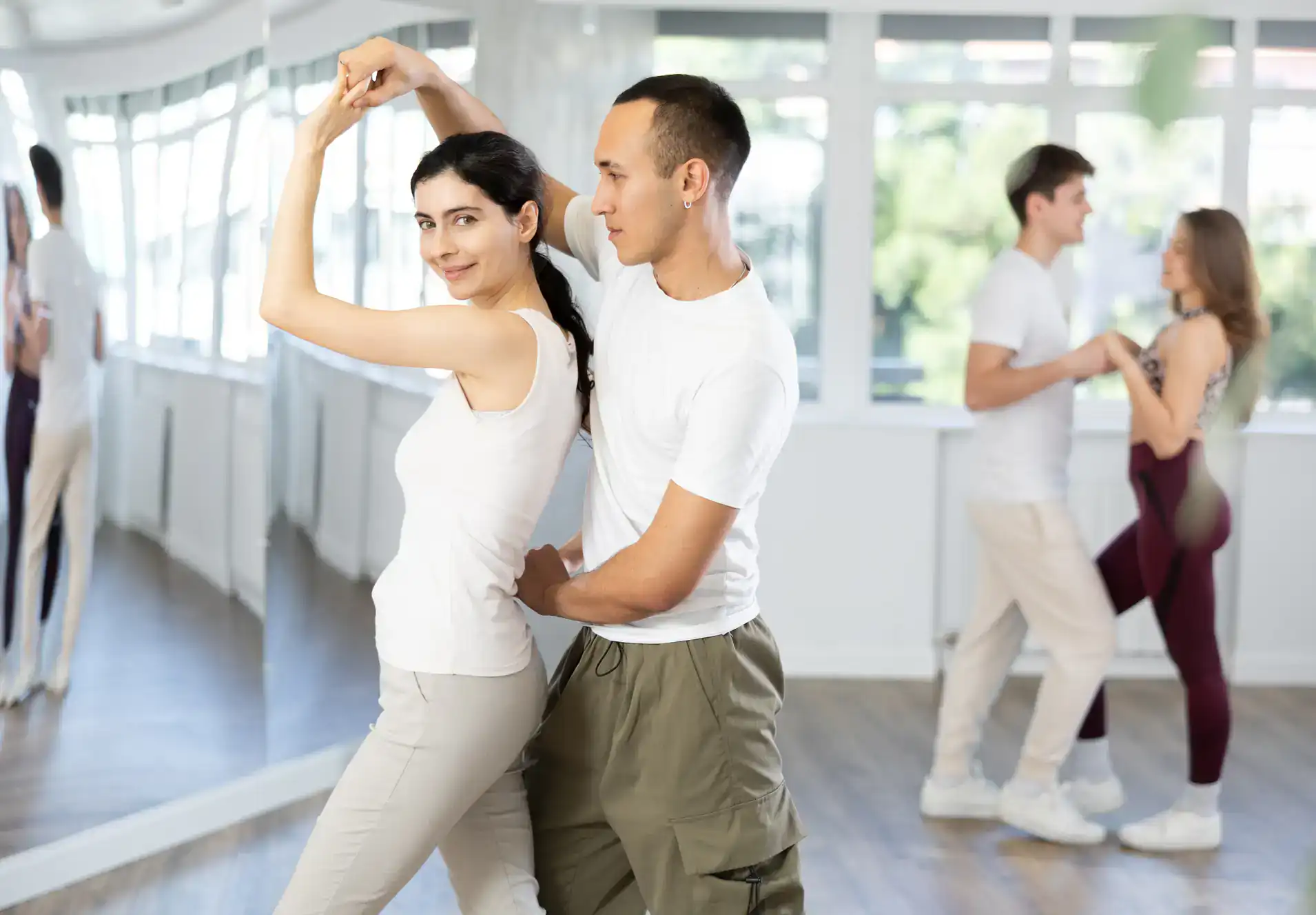 Two couples practice ballroom dance Suffolk County in a bright studio with large windows. The foreground couple poses in a dance hold, while the background couple dances together, both wearing casual clothes.