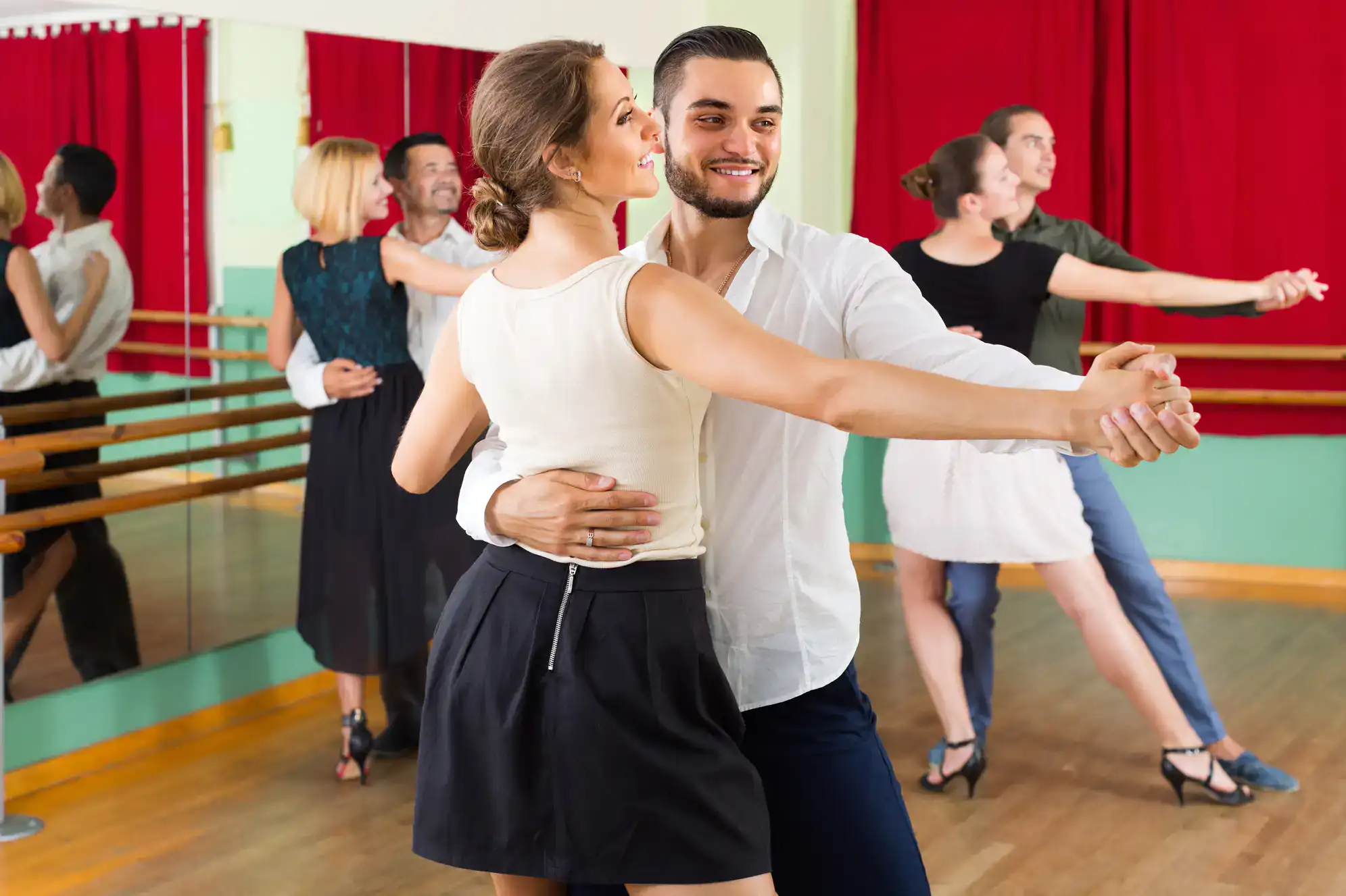 Four couples practice ballroom dance in a brightly lit NY studio with wooden floors, mirrors, and red curtains. The focus is on a smiling pair at the front holding a pose—showcasing Dance Lessons Suffolk County style.