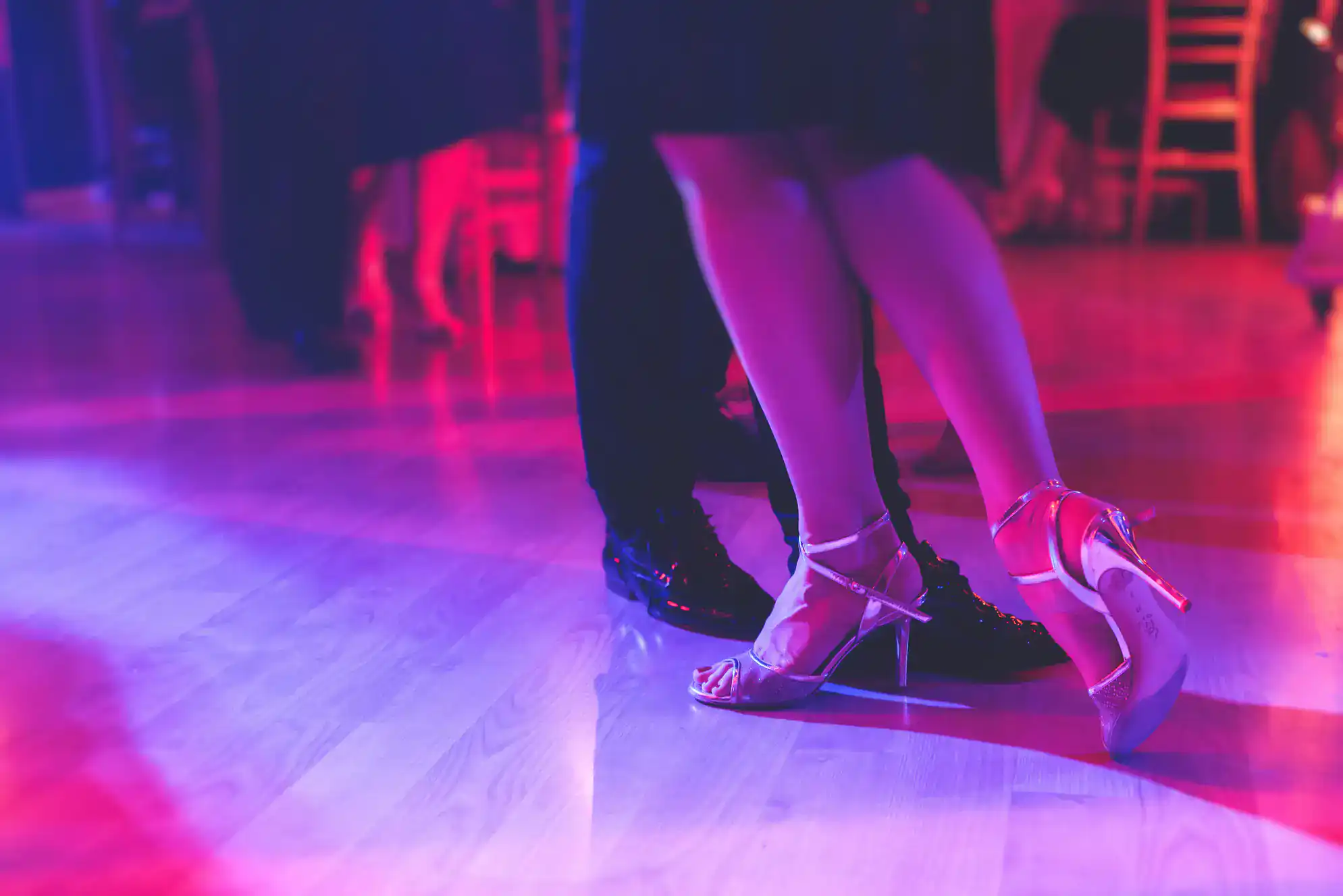 A close-up of two people&rsquo;s legs dancing together on a wooden floor, one in high-heeled sandals and the other in black dress shoes, under pink and purple lighting&mdash;capturing the elegance of ballroom dance Suffolk County, NY.