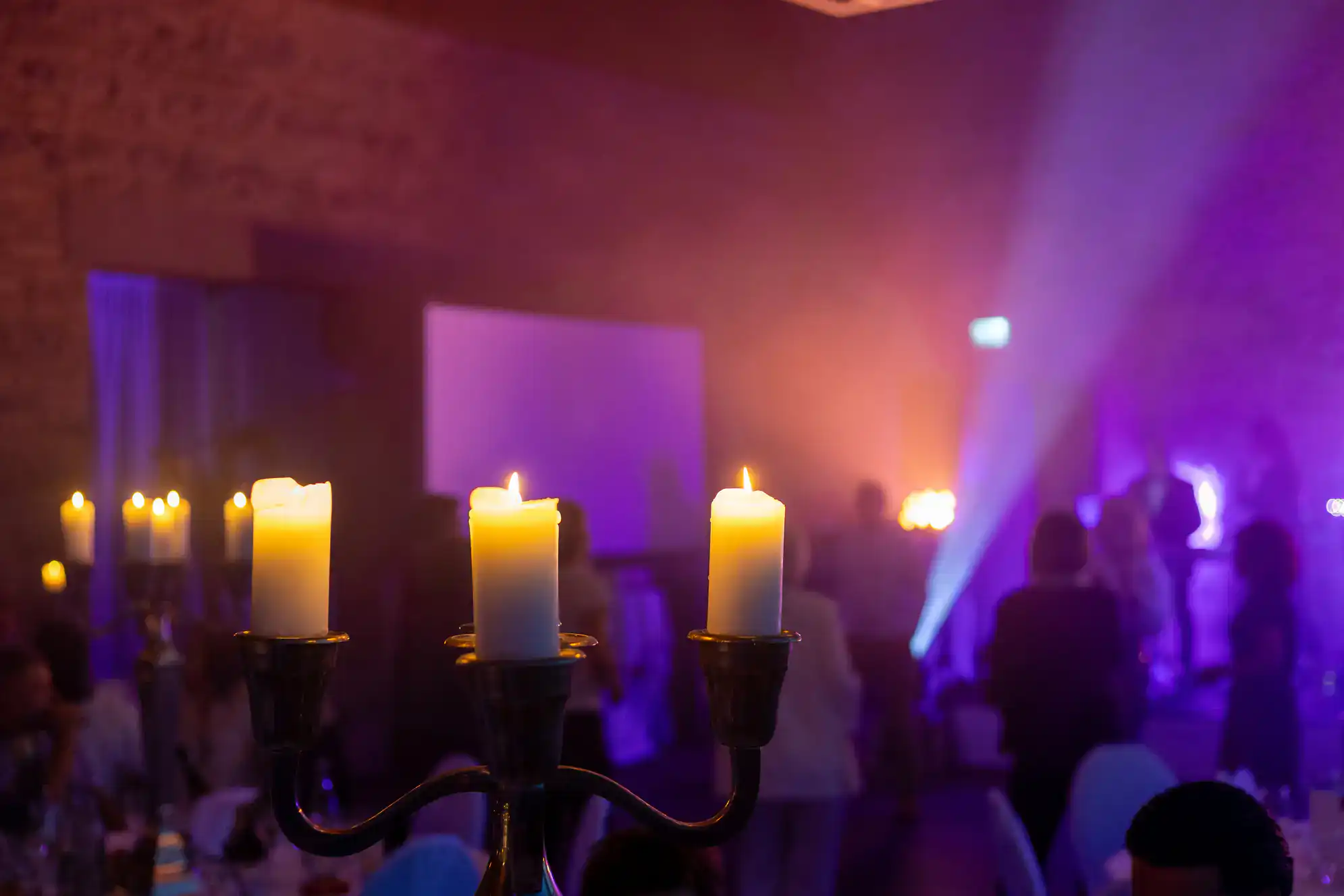 A close-up of lit candles on a candelabra in the foreground with a blurred background of people and colorful lights at a dimly lit NY indoor event, perfect for ballroom dance Suffolk County nights.