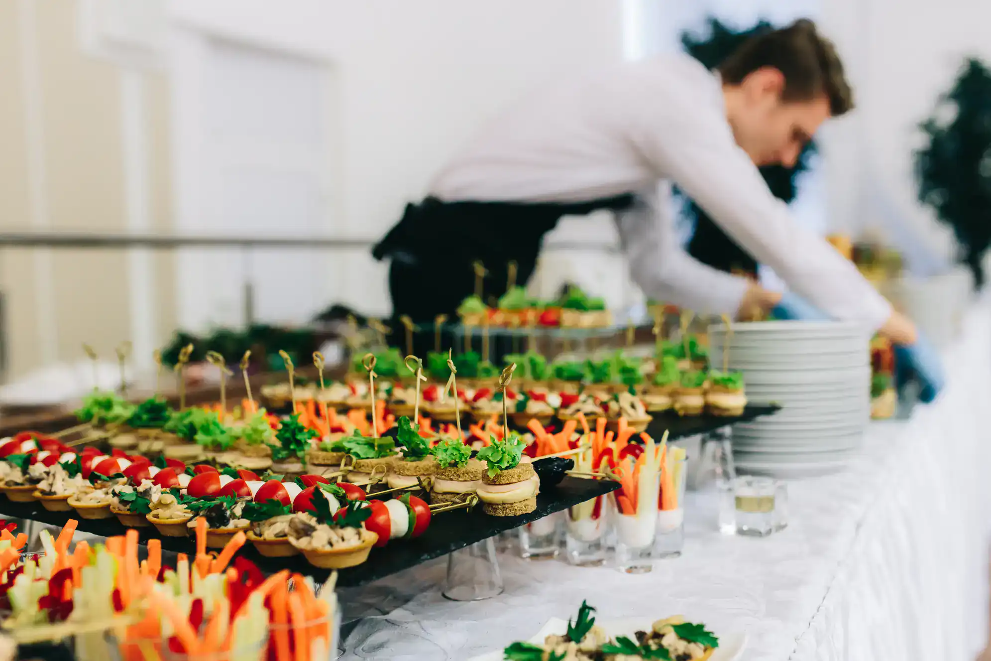 A person in a white shirt and black apron arranges plates behind a table filled with assorted appetizers, including vegetable cups, tartlets, and skewers, at a NY catered event featuring ballroom dance Suffolk County.
