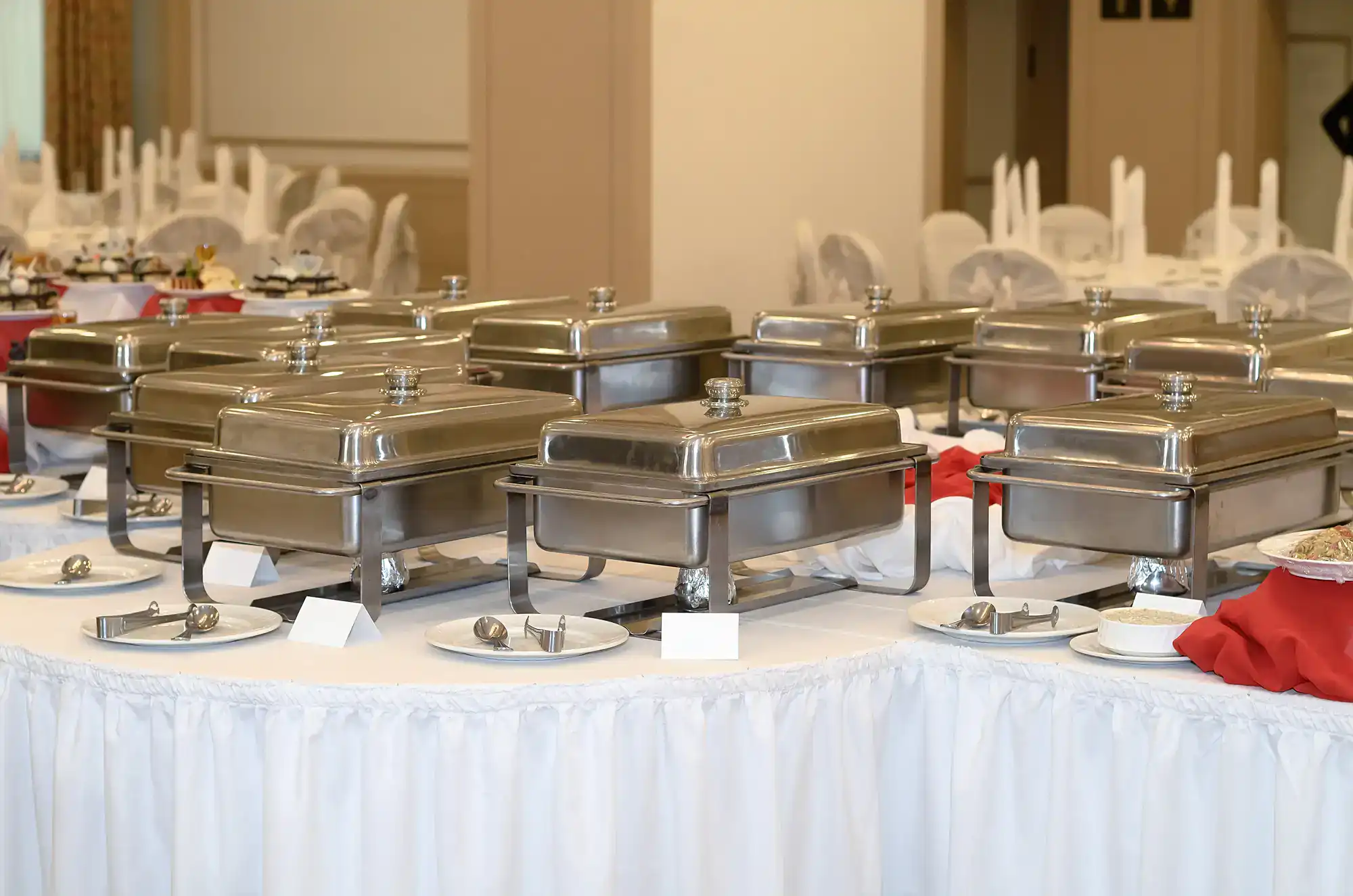 A banquet setup with several closed stainless steel chafing dishes on white-clothed tables, surrounded by plates and serving utensils, ready for a buffet service in a decorated NY dining hall after ballroom dance Suffolk County lessons.