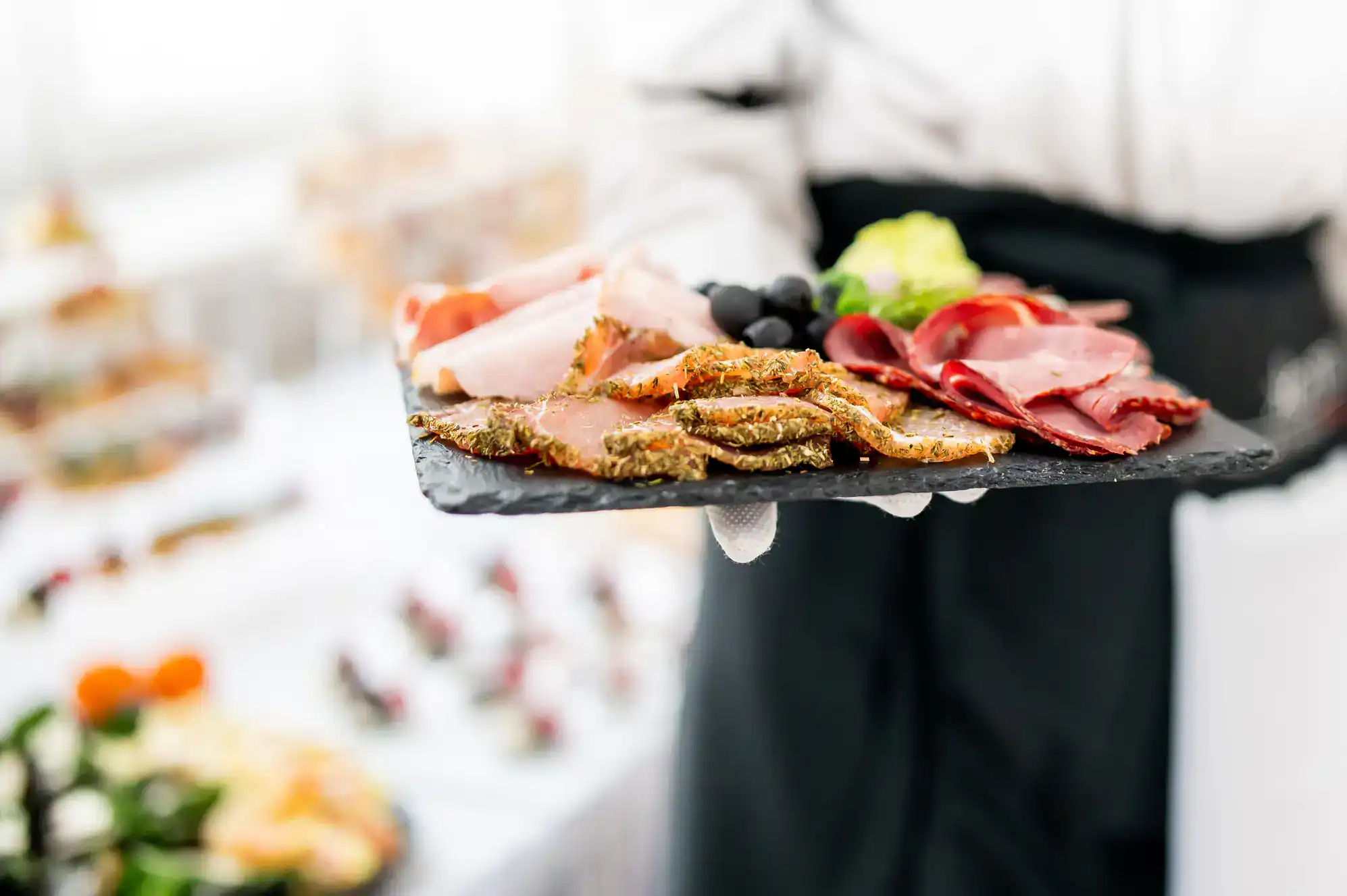 A server wearing white gloves presents a slate platter with assorted cured meats, fresh greens, and dark grapes&mdash;perfect for savoring after ballroom dance lessons in Suffolk County, NY. A blurred table of various foods completes the elegant scene.