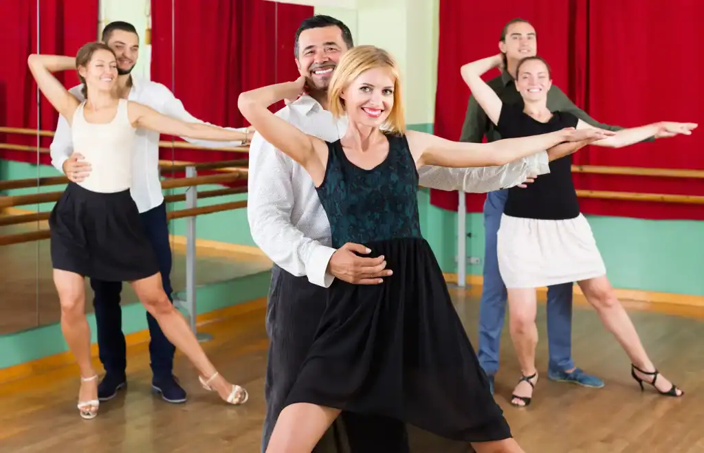 Three smiling couples pose in a dance studio with wooden floors and mirrors in Suffolk County, NY. The front couple holds a ballroom dance position, showcasing the elegance learned from Dance Lessons Suffolk County, NY offers.
