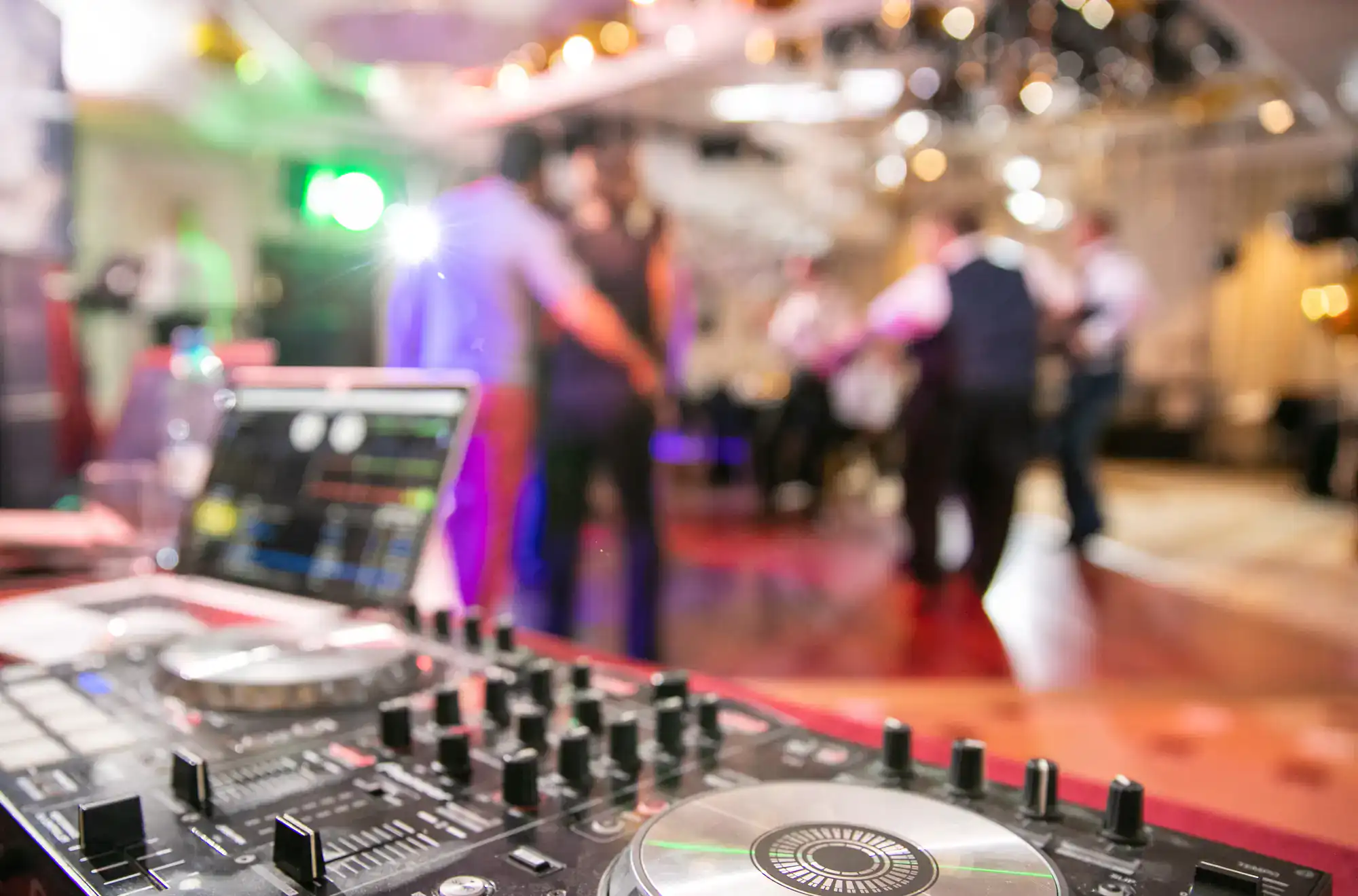 Close-up of a DJ mixing console with a laptop in the foreground, while people in formal attire enjoy ballroom dance in Suffolk County on a colorful, blurred dance floor. Bright lights complete the festive NY atmosphere.
