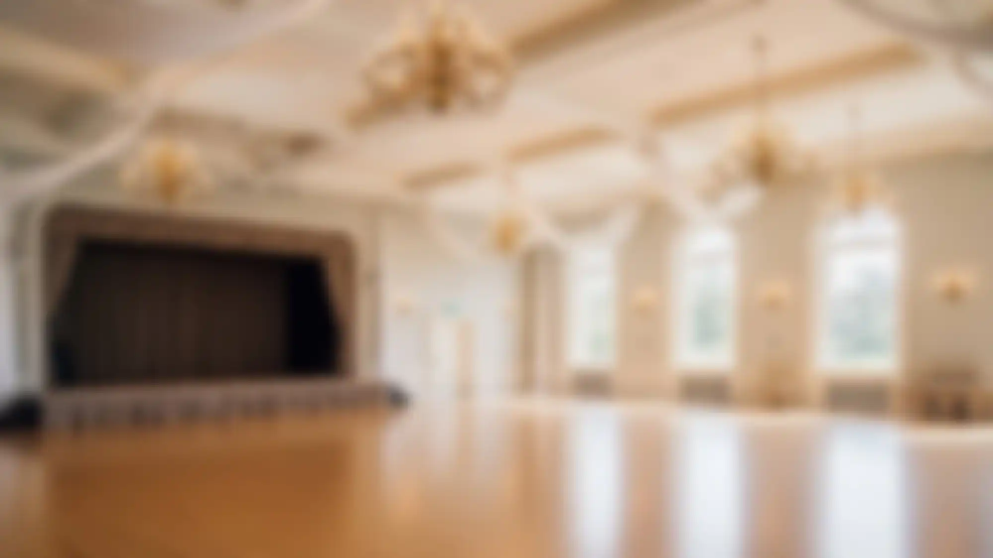 A large, elegant NY ballroom with high ceilings, chandeliers, tall windows, and a stage at one end&mdash;perfect for ballroom dance Suffolk County events. The empty room is well-lit, with wooden floors and light walls. The image is blurred.