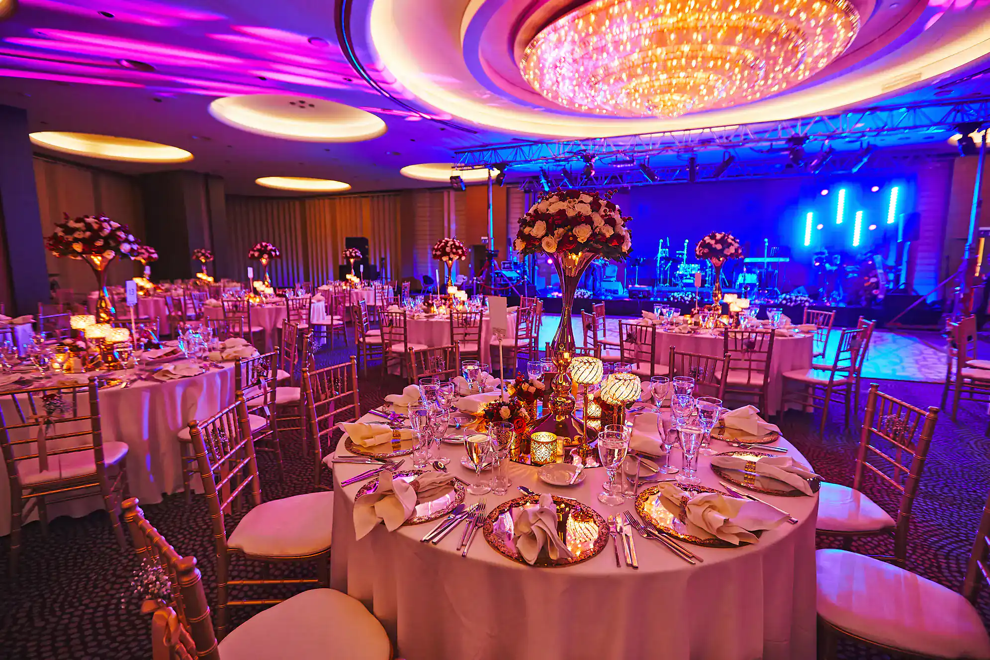 A luxurious banquet hall in NY set for an event, featuring round tables with elegant place settings, floral centerpieces, glowing candles, and gold chairs&mdash;perfect for ballroom dance Suffolk County&mdash;under a large, sparkling chandelier with vibrant purple lighting.