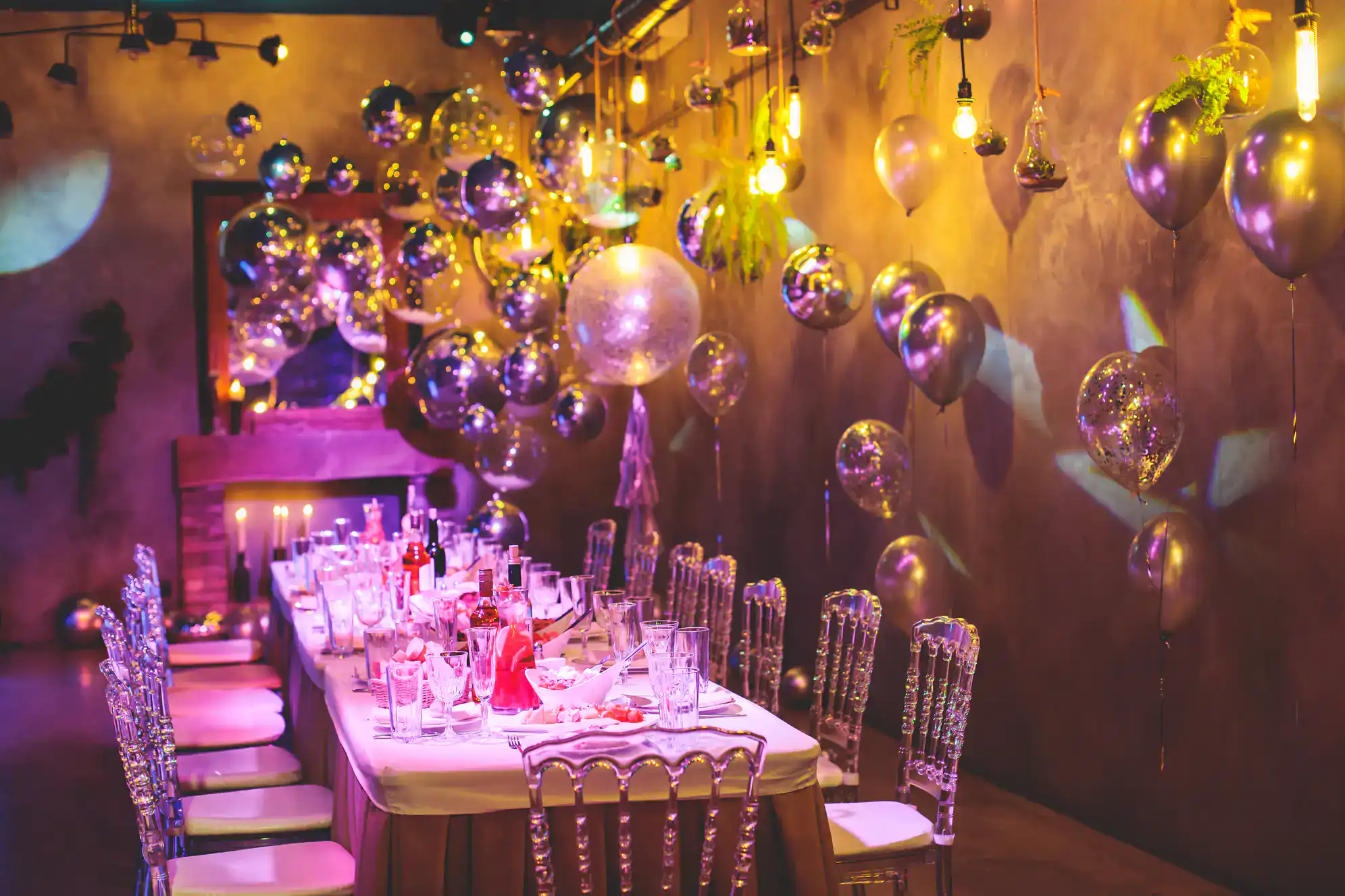 A long dining table is set for a party with elegant chairs, glassware, and candles. The room, perfect for ballroom dance Suffolk County events, features metallic balloons and warm hanging lights for a festive, glamorous NY atmosphere.