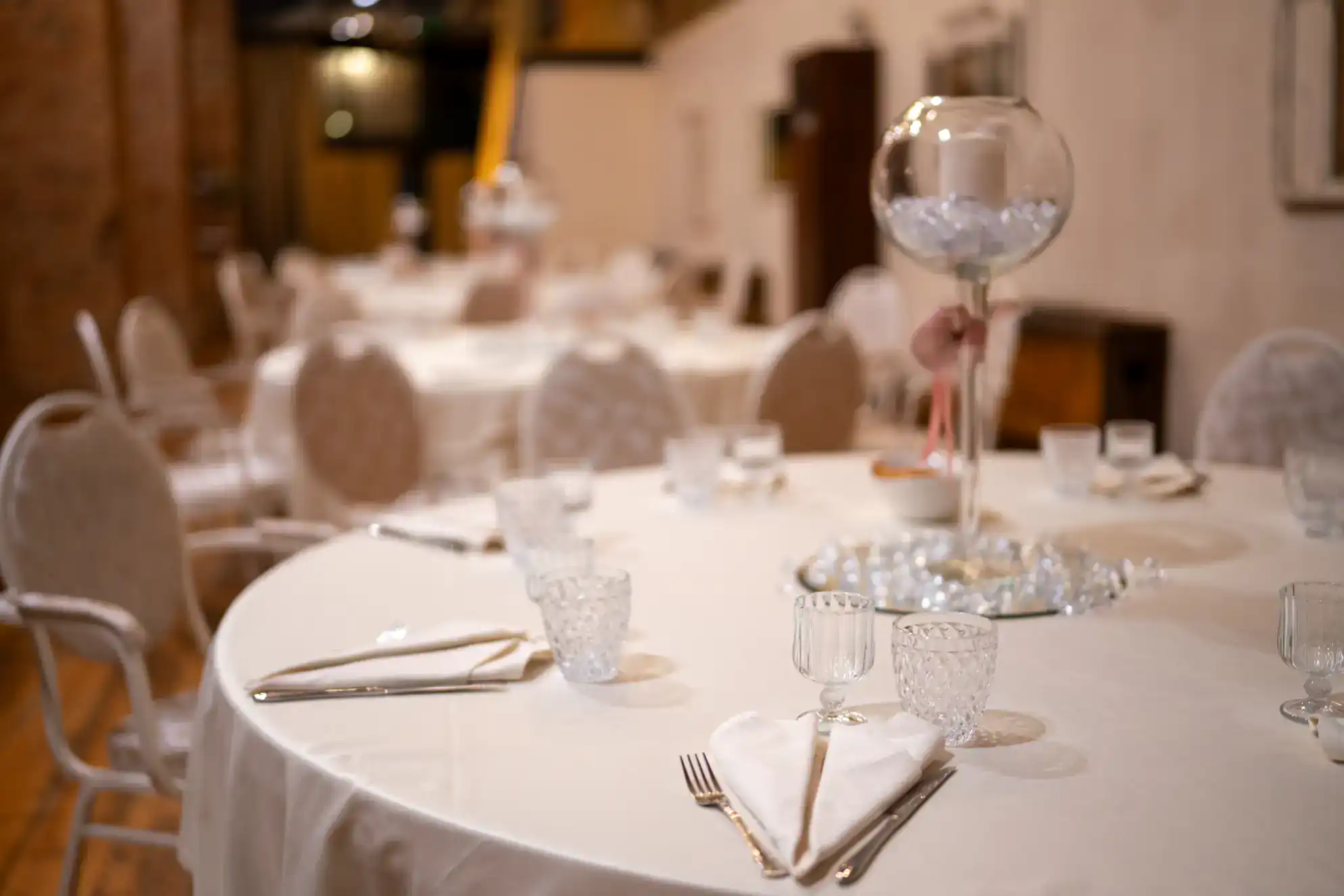 A round banquet table is elegantly set with glassware, plates, and white napkins in a softly lit dining room&mdash;perfect for celebrating after ballroom dance lessons in Suffolk County, NY. A large decorative glass centerpiece sits on top.