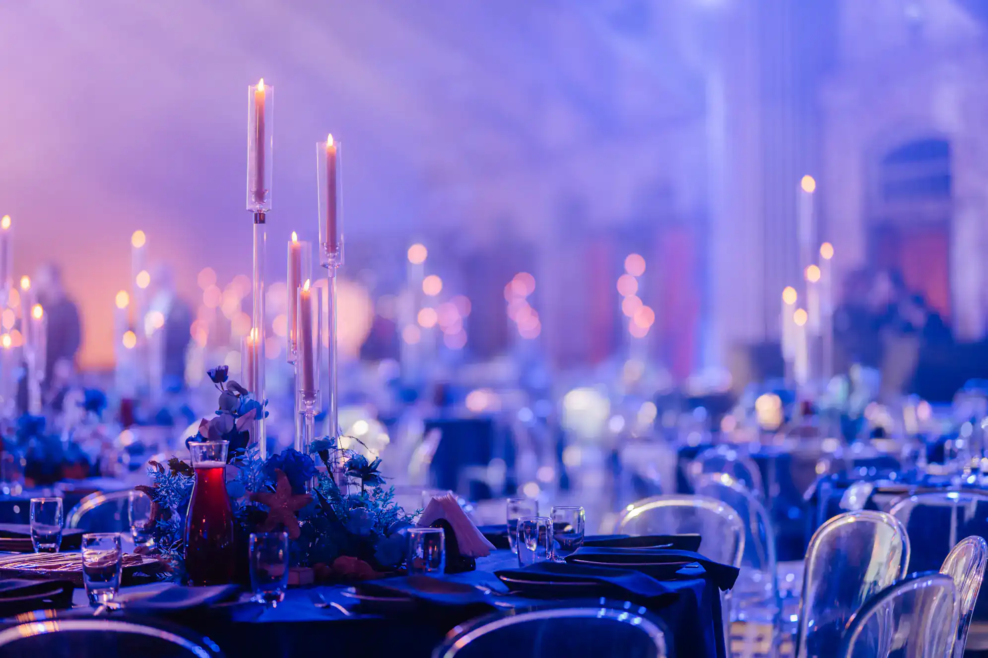 A formal banquet hall with elegantly set tables, tall lit candles in glass holders, floral centerpieces, and clear chairs. Soft blue lighting creates a festive and sophisticated atmosphere&mdash;perfect for ballroom dance Suffolk County, NY events.