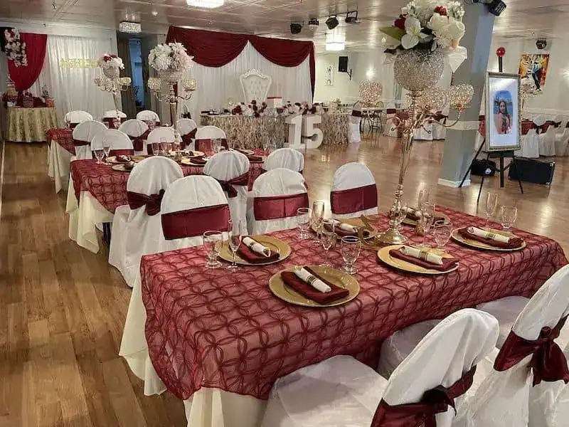 Decorated event hall in NY with tables covered in red-patterned cloths, gold charger plates, and white chair covers with red sashes&mdash;perfect for ballroom dance Suffolk County celebrations. Tall floral centerpieces and elegant place settings adorn every table.