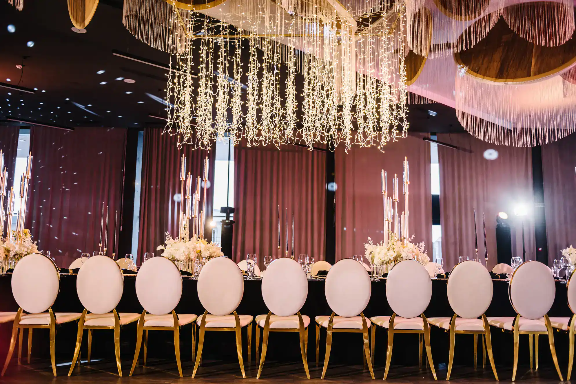 Elegant, formal dining table with gold and white chairs, elaborate floral centerpieces, tall candles, and a sparkling chandelier of string lights&mdash;perfect for a glamorous ballroom dance Suffolk County, NY event in a luxurious venue with dark curtains.