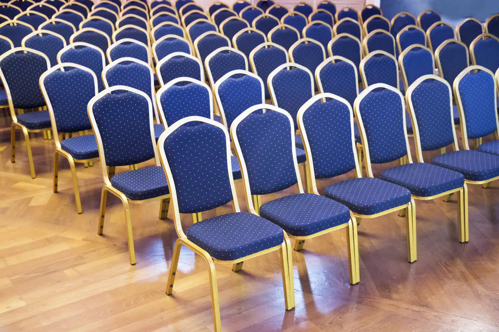 Rows of empty blue cushioned chairs with gold frames are arranged on a wooden floor, facing the same direction in what appears to be a ballroom dance Suffolk County, NY event hall.