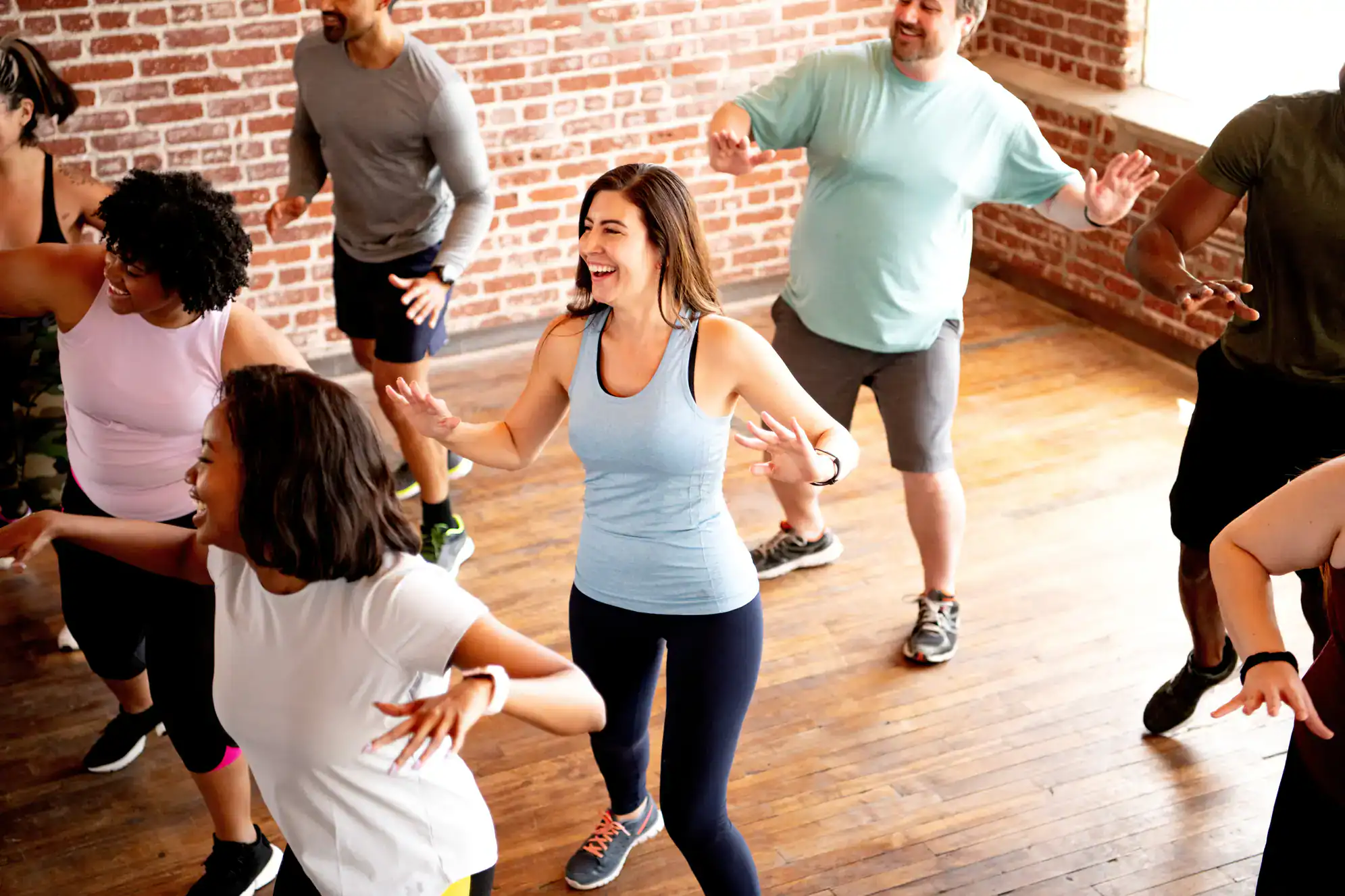 A group of people exercise together in a studio with wooden floors and brick walls, smiling and actively participating in a dance or fitness class. Experience fun Dance Lessons Suffolk County, NY has to offer while wearing casual athletic clothing.