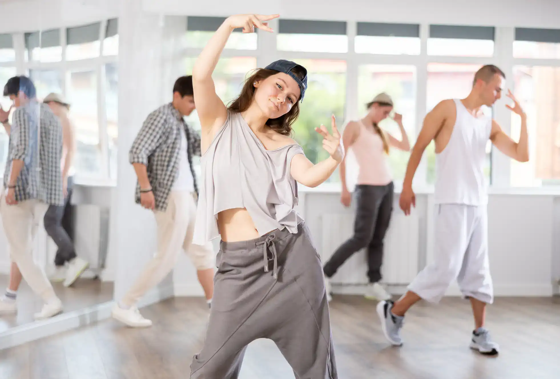A young woman in loose clothing and a backwards cap poses confidently in a NY dance studio, while three others practice ballroom dance Suffolk County style near a large mirror.