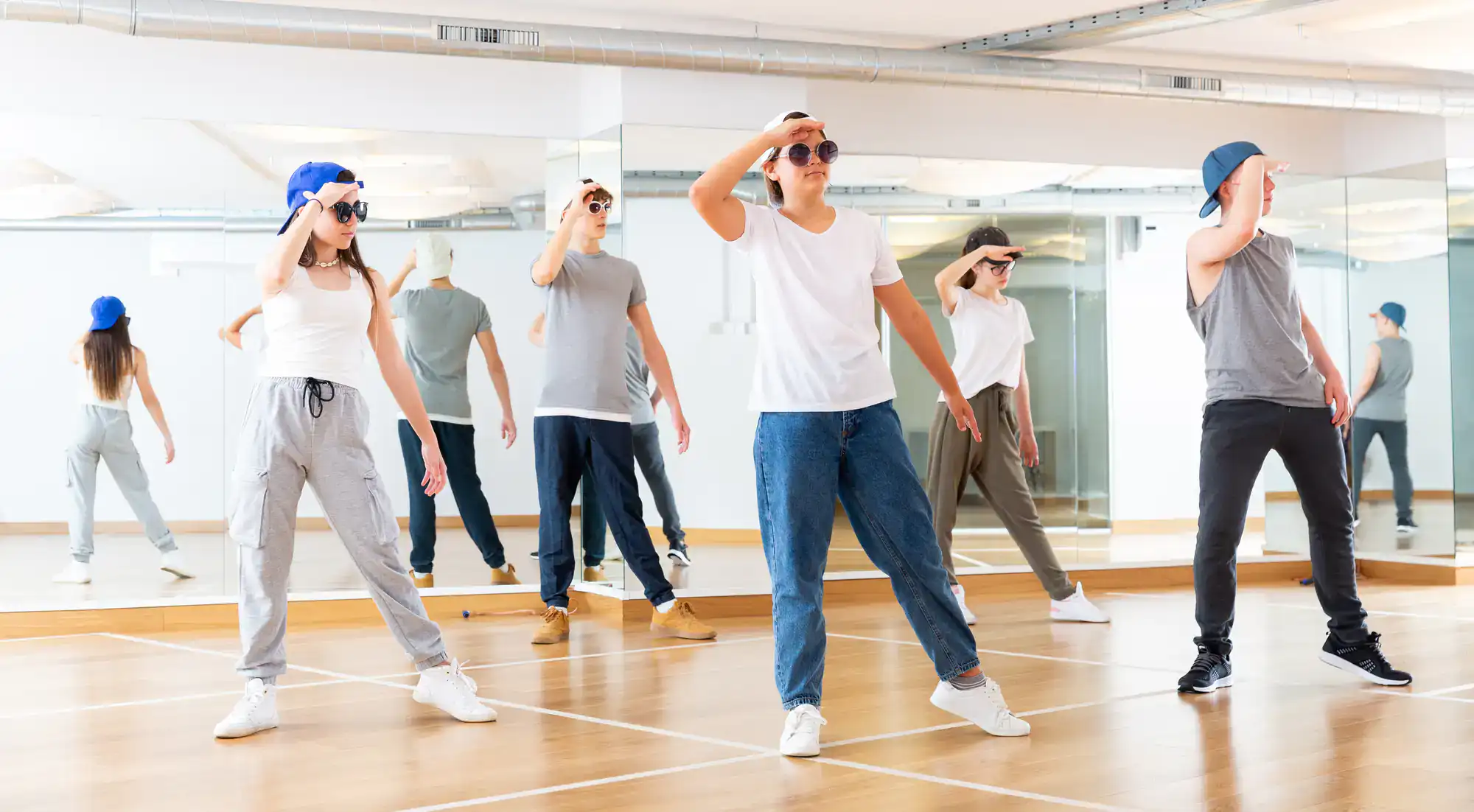 A group of people in casual clothes and hats practice a dance routine in a mirrored studio, all striking a pose with one hand on their foreheads as if saluting during ballroom dance lessons Suffolk County, NY.