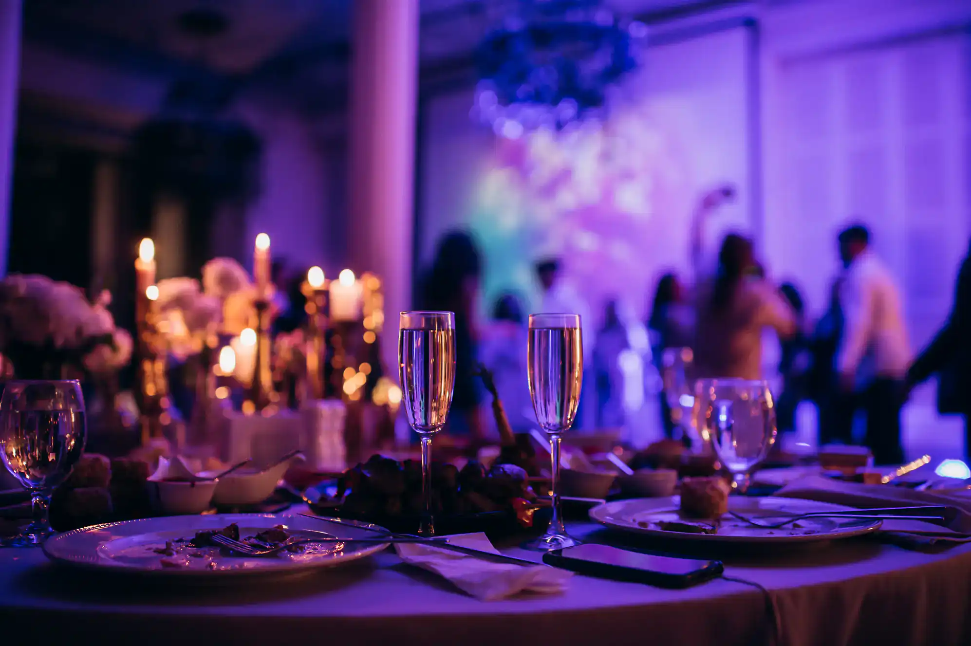 A dimly lit banquet table set with plates, food, and two glasses of champagne. In the background, people are mingling under purple lighting, creating a festive atmosphere inspired by ballroom dance Suffolk County events.