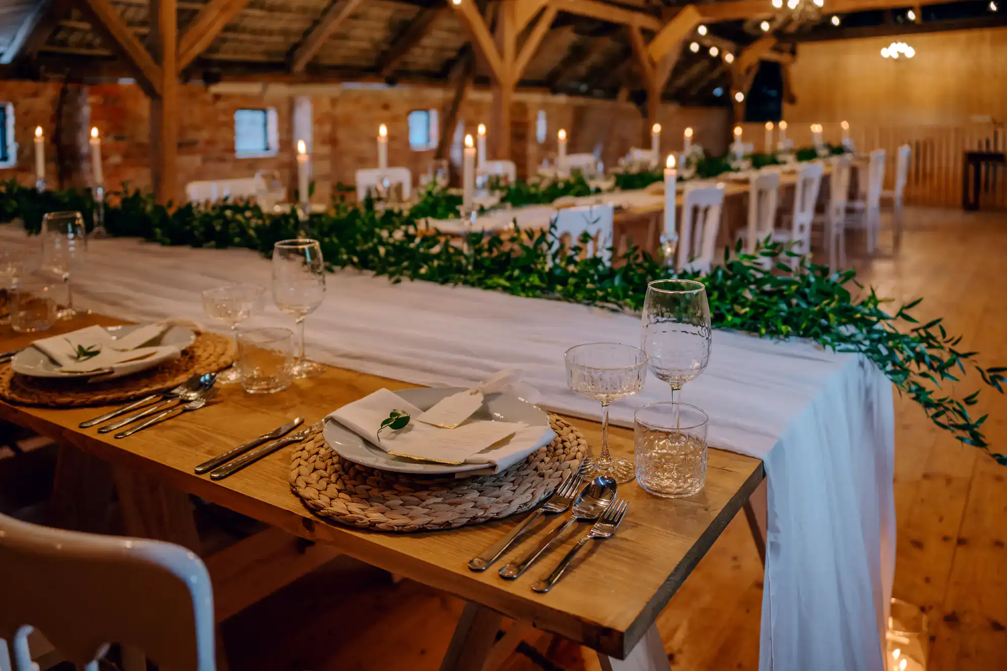 Elegant rustic table setting in a wooden NY venue with long tables, white plates, glassware, silver cutlery, woven placemats, green garland centerpieces, white candles, and soft warm lighting&mdash;perfect for a ballroom dance Suffolk County event.