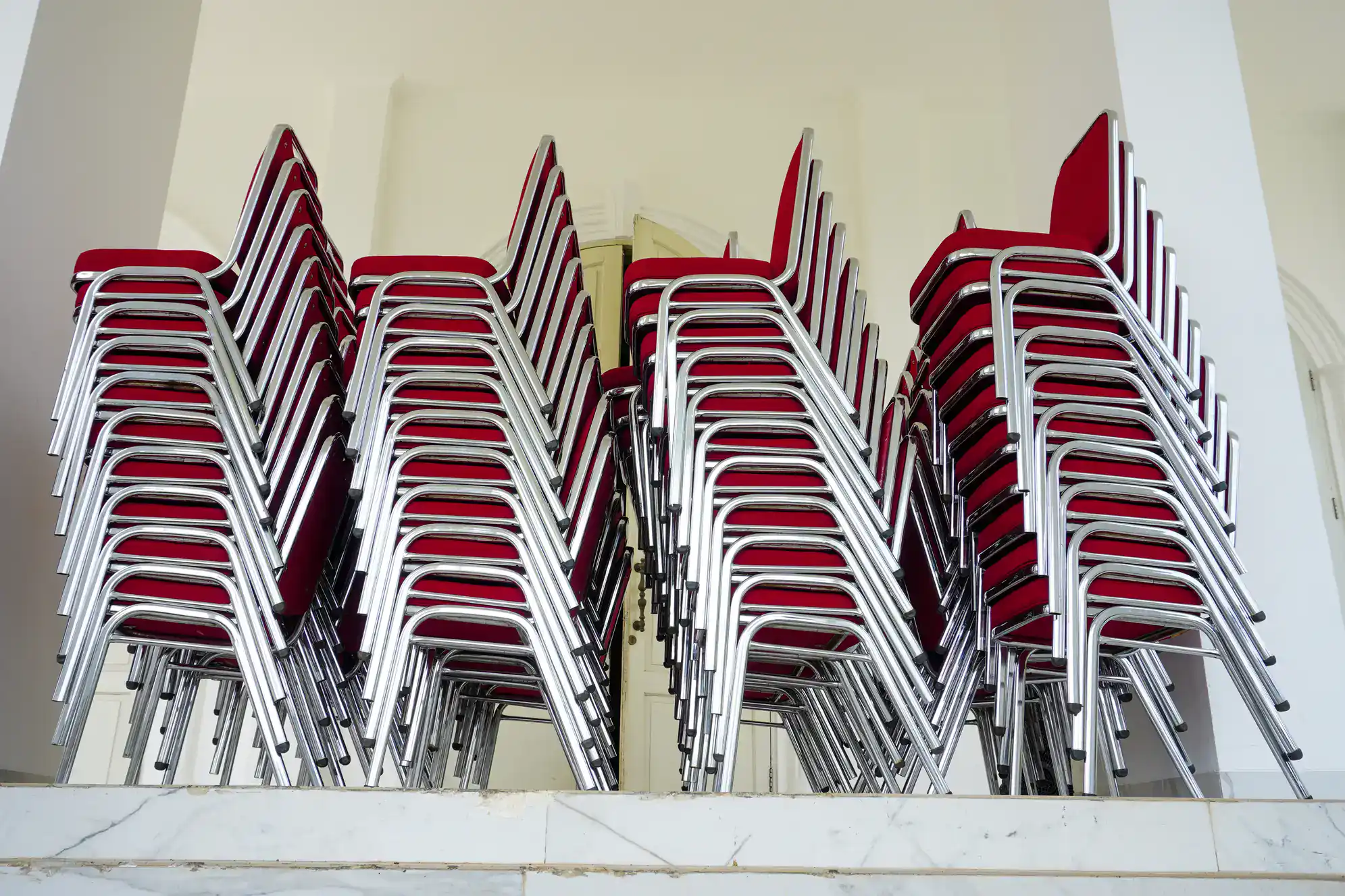 Several tall stacks of metal chairs with red seats and backs are arranged closely together on a marble platform in a bright, white room&mdash;perfect for hosting NY ballroom dance Suffolk County events or Dance Lessons Suffolk County.
