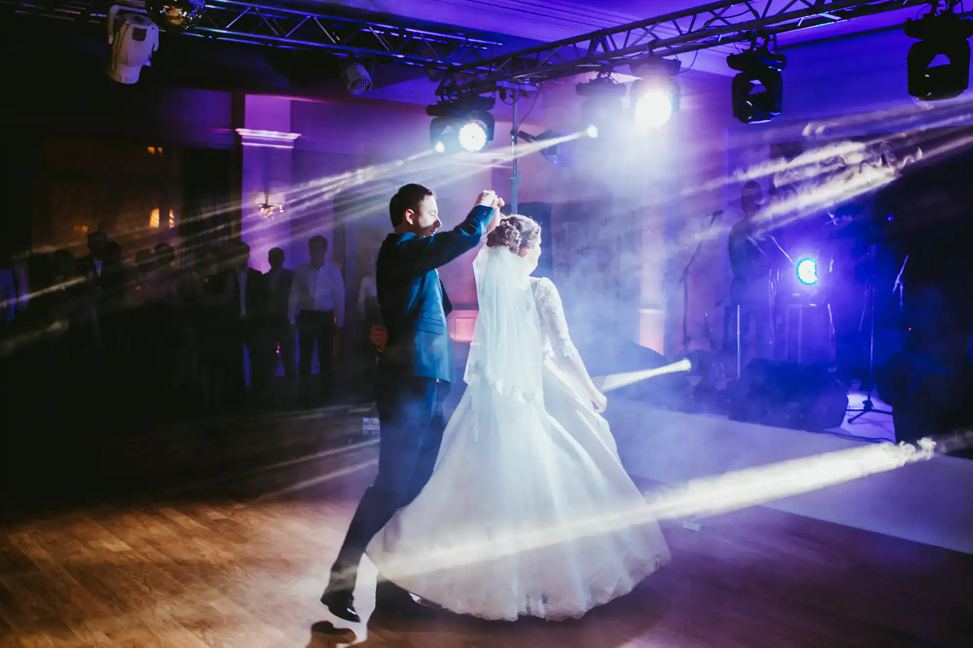 A bride and groom, having taken dance lessons in Suffolk County, share a romantic first dance under dramatic stage lighting as beams of light shine around them in a dimly lit wedding reception hall. Guests watch in the background.