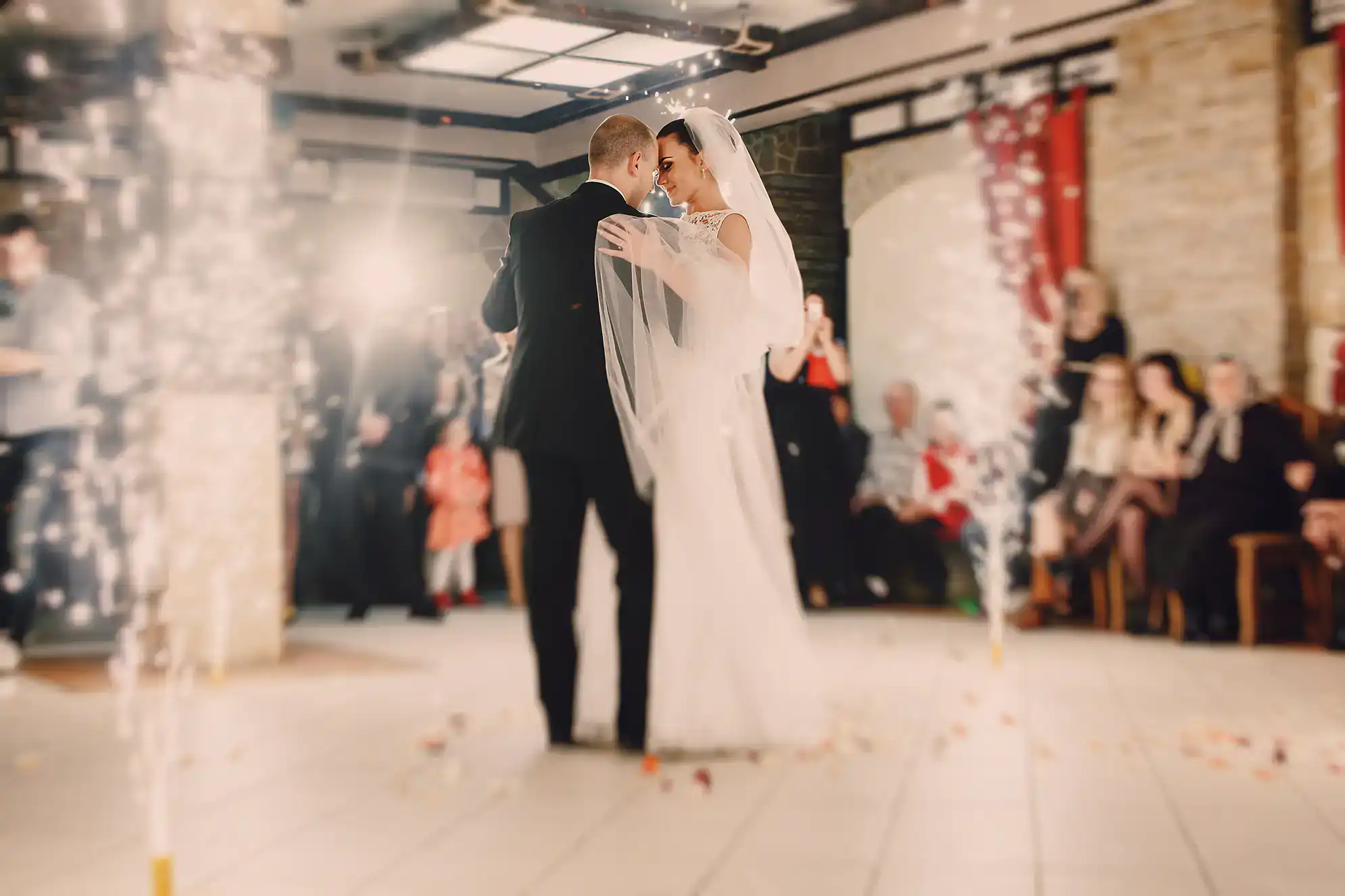 A bride and groom share their first dance at their wedding reception, surrounded by guests and indoor fireworks, with soft lighting creating a romantic atmosphere&mdash;thanks to ballroom dance lessons in Suffolk County, NY.