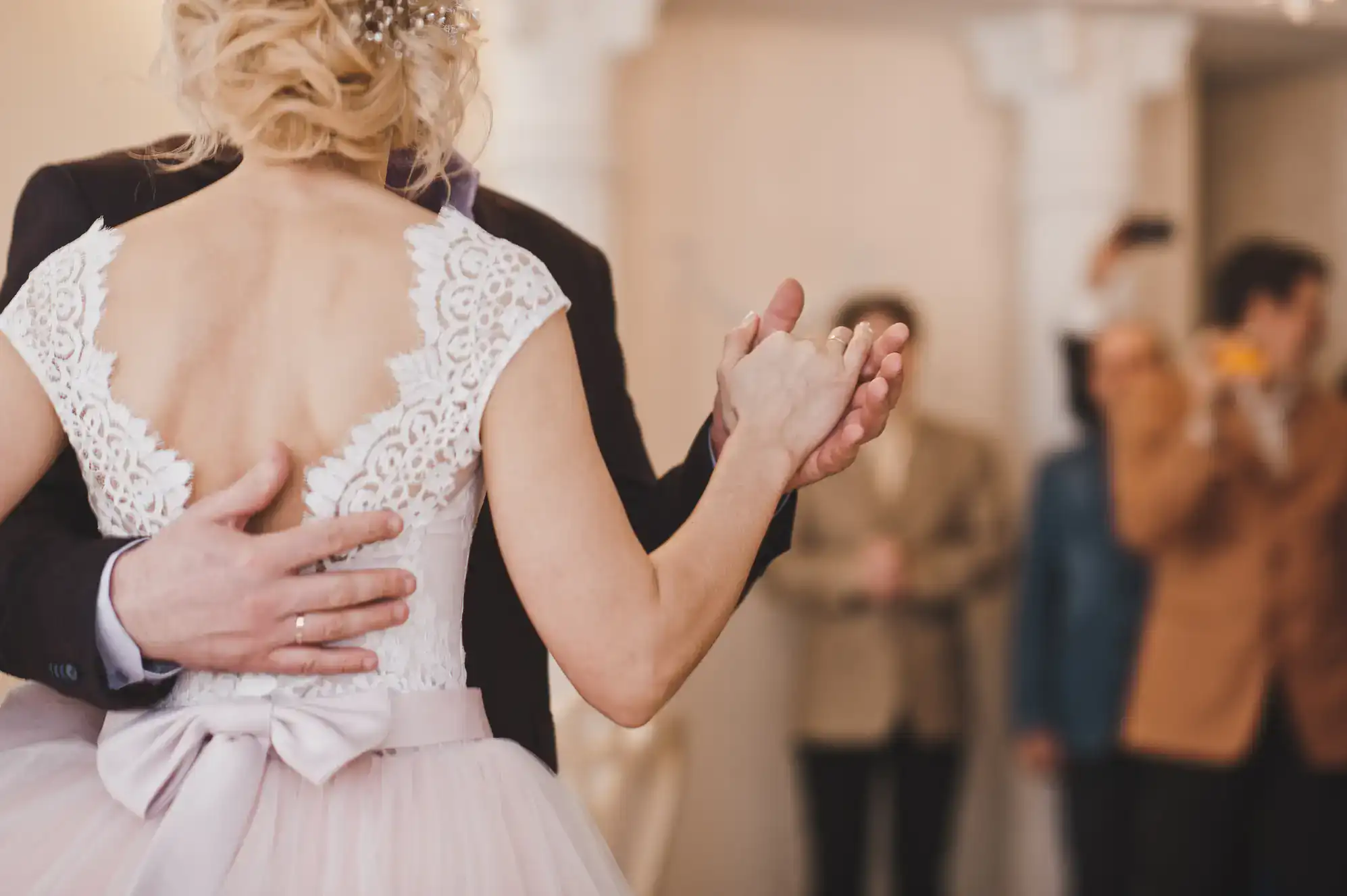 A bride in a white lace dress and groom in a suit, having taken ballroom dance Suffolk County lessons, dance closely while holding hands. The bride's back faces the camera as blurred guests in the background watch and take photos.