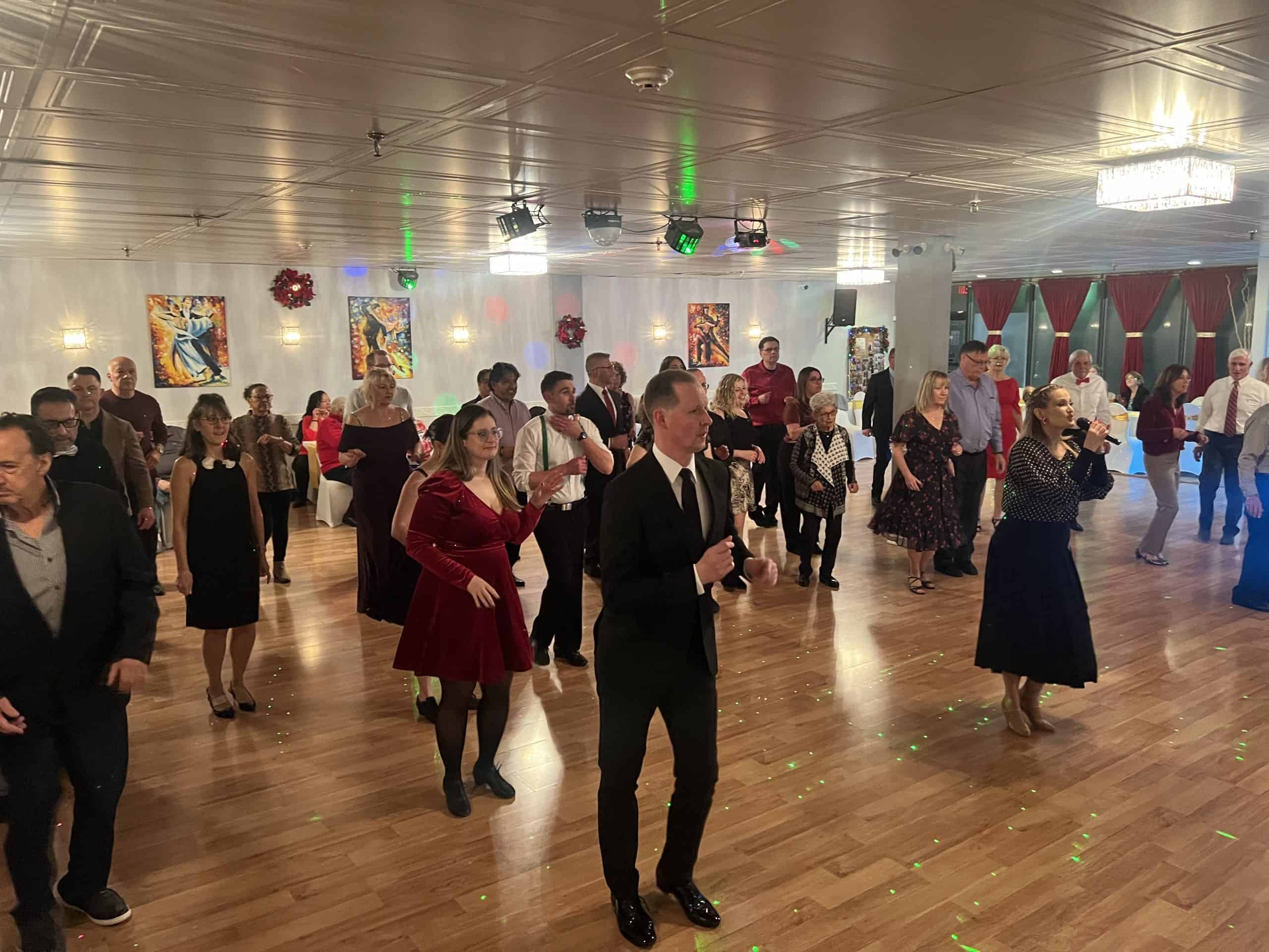 People in formal attire dance in a well-lit ballroom with art, festive holiday decor, and elegant lighting.